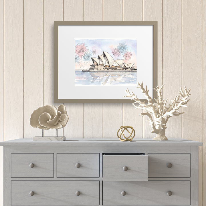 A framed art print depicting the Sydney Opera House with fireworks and a sailboat, styled on a light gray wooden dresser against a beige wood-paneled wall.