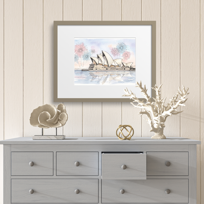 A framed art print depicting the Sydney Opera House with fireworks and a sailboat, styled on a light gray wooden dresser against a beige wood-paneled wall.