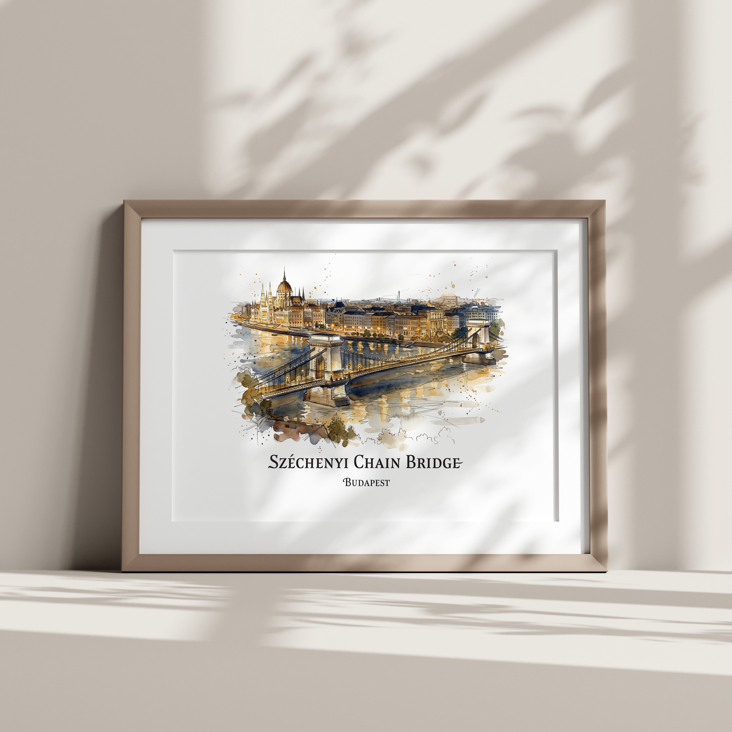 A framed watercolour art print of the Széchenyi Chain Bridge, Budapest 
displayed on a shelf against a wall with soft leaf shadows.