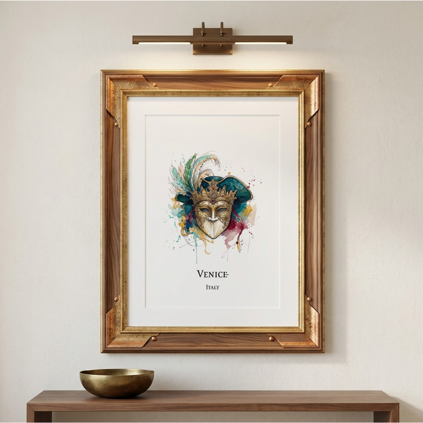 A framed art print featuring a colourful watercolour Venetian mask with the text 'VENICE' and 'ITALY' below, displayed on a beige wall above a wooden console table with a gold bowl. A brass wall sconce is mounted above the frame.