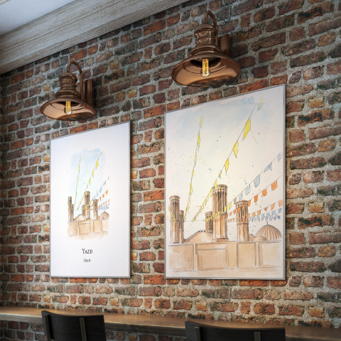 Two vertical framed art prints with watercolour illustrations of Yazd in Iran, hanging on a rustic red-brown brick wall in a cafe or restaurant setting, with two industrial-style copper wall sconces above them.