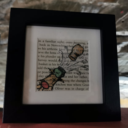 A freestanding black 12.5 cm box frame on a bookshelf against a back lit slate wall. An internal 6cm frame has a book page with a hand-drawn watercolour illustration of a champagne bottle pointing up to the right with the cork popping.