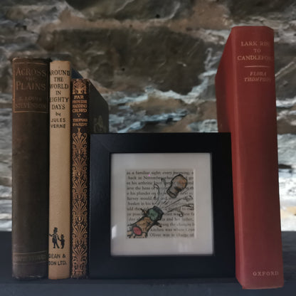 A freestanding black 12.5 cm box frame on a bookshelf against a slate wall with vintage books. An internal 6cm frame has a book page with a hand-drawn watercolour illustration of a champagne bottle pointing up to the right with the cork popping.